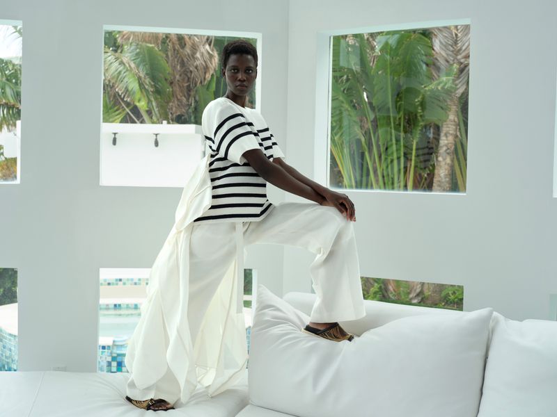 A woman with dark skin in an off-white striped top and white pants poses indoors against a white wall with square windows.