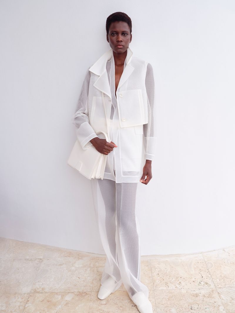 A confident Black woman poses in an all-white semi-sheer fashion outfit against a minimalist white wall, showcasing style and elegance.