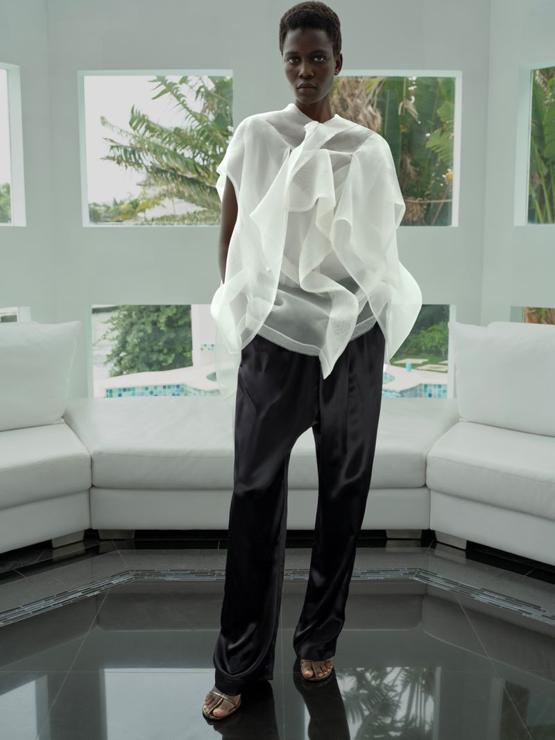 Fashion shot of a slender woman in a white sheer top and black wide-legged pants posing indoors near white sofa and large windows.