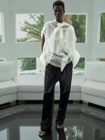 Fashion shot of a slender woman in a white sheer top and black wide-legged pants posing indoors near white sofa and large windows.