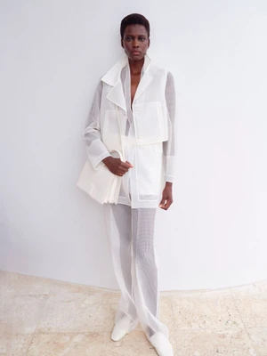 Model standing against a neutral background wearing a sheer white layered outfit and holding a white handbag.