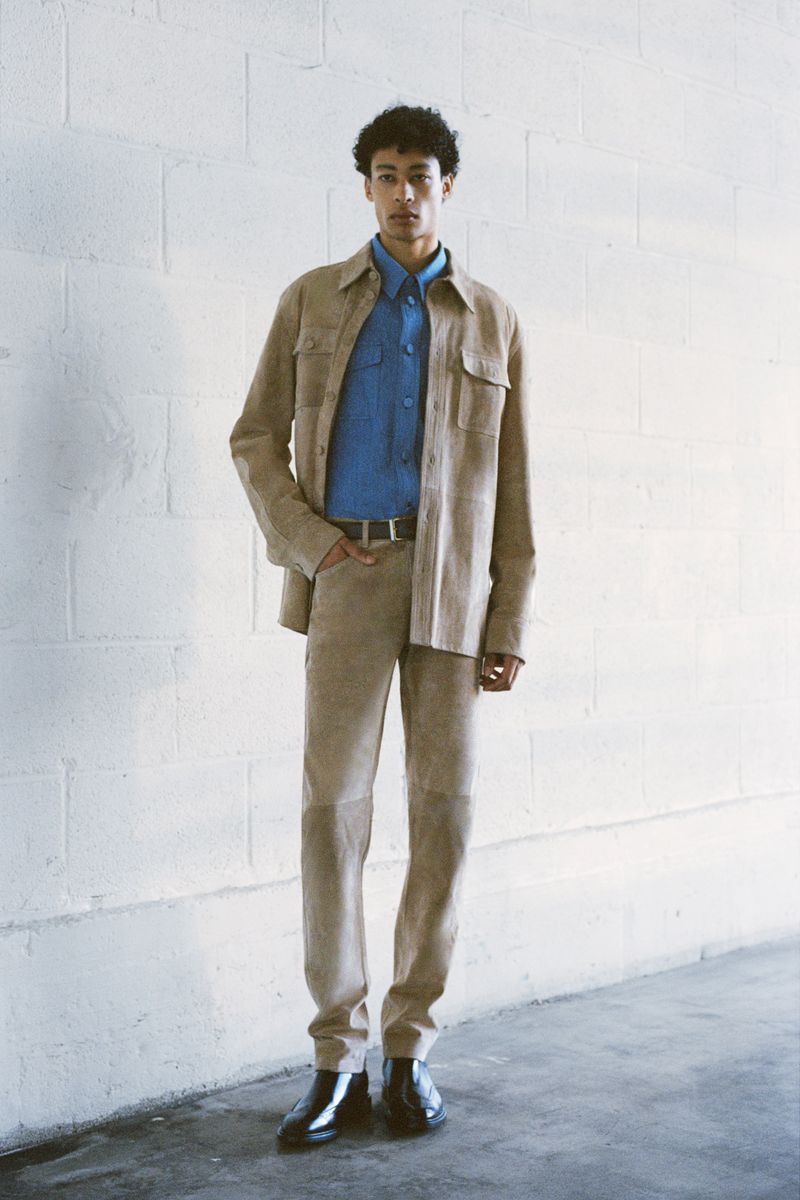 A curly-haired man in a light brown suede outfit and black boots stands against a white brick wall in a fashion style photograph.