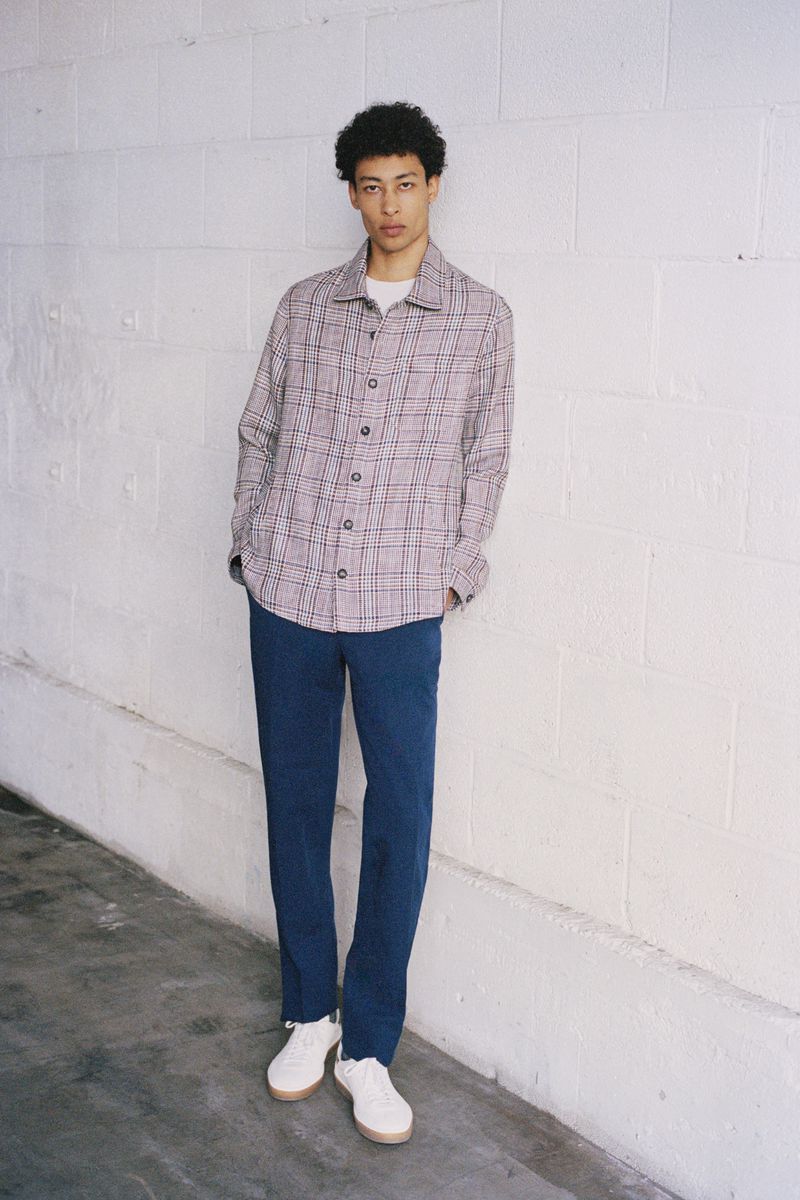 A stylish young man poses in a plaid shirt and blue pants, standing against a textured white wall, showcasing a casual fashion ensemble.