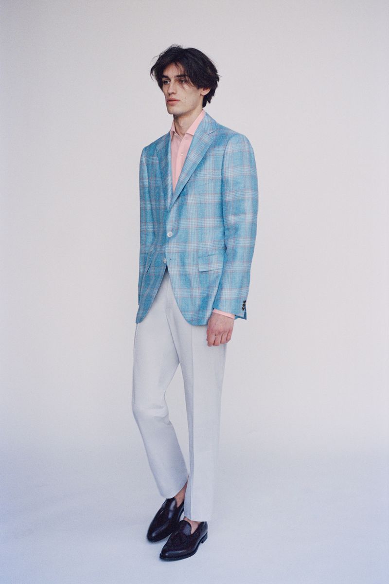 Model wearing a blue plaid blazer, pink shirt, and white trousers standing against a white seamless backdrop