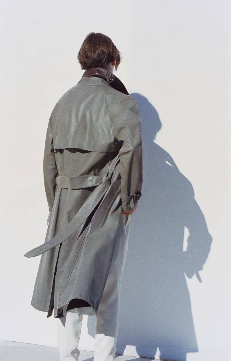Person from behind wearing a grey leather trench coat, white pants, standing in front of a white wall with their shadow visible.