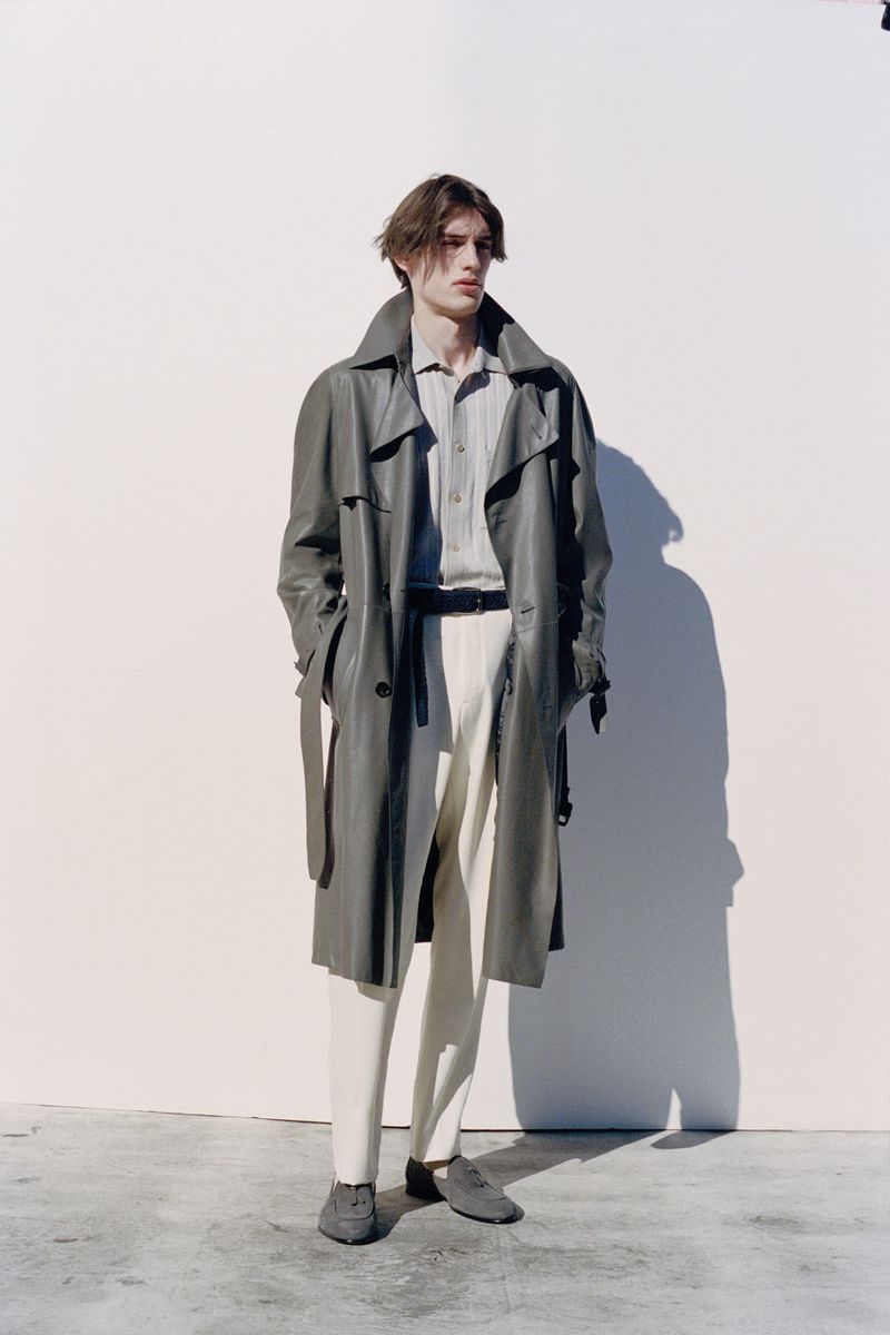 A fashionable male model poses against a white wall, wearing a long trench coat, striped shirt, white pants, and loafers in an outdoor setting.