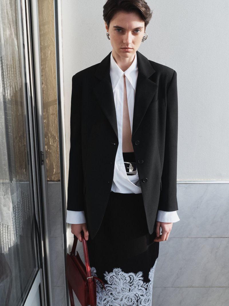 Adult woman in a black blazer and black skirt with lace trim carries a red bag in an indoor setting with natural lighting.