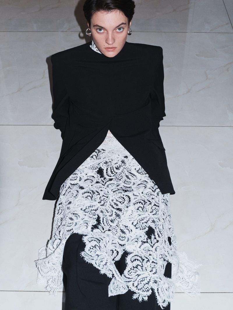 A woman in a black jacket and white lace skirt stands indoors against a neutral wall, looking at the camera in a fashion shot.