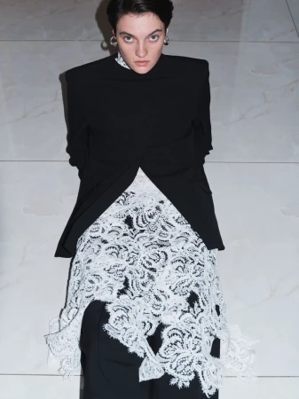 A woman in a black jacket and white lace skirt stands indoors against a neutral wall, looking at the camera in a fashion shot.
