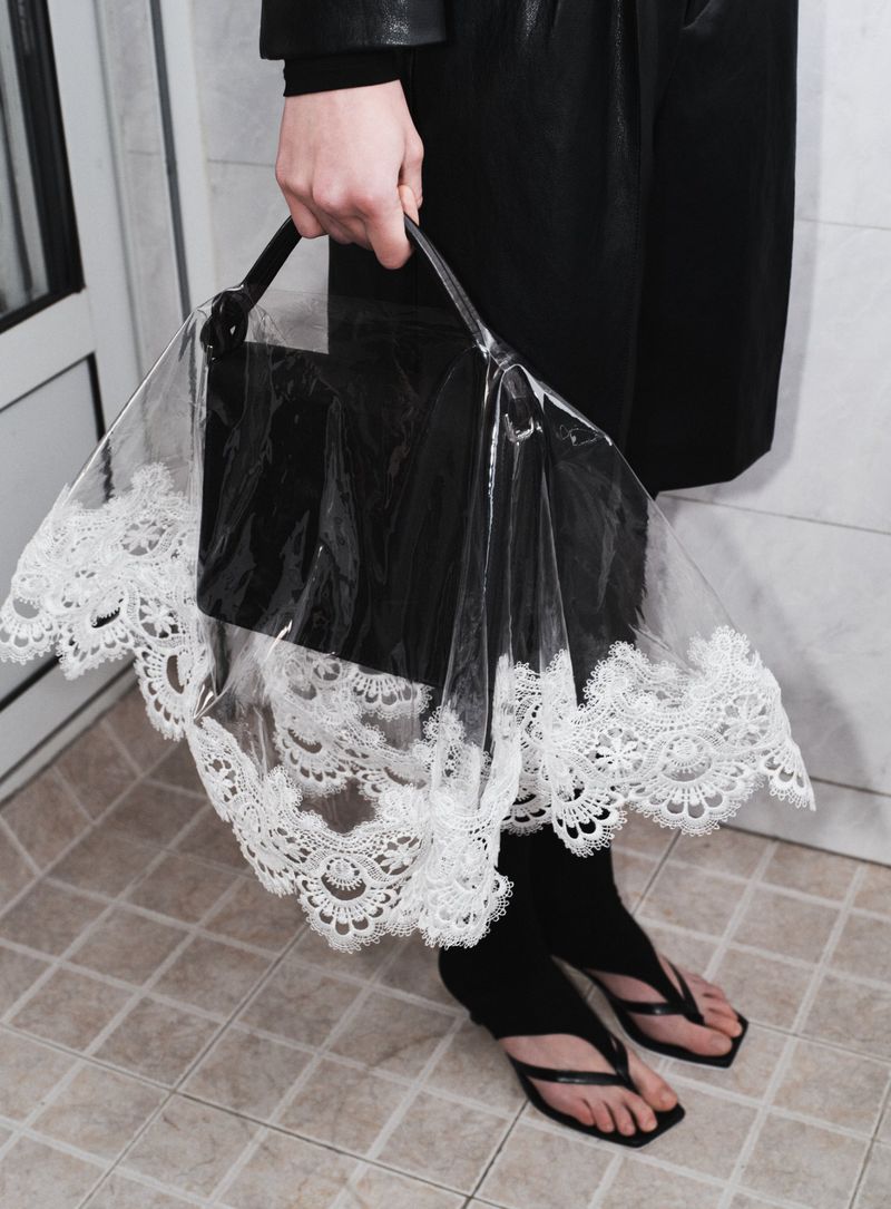 Close-up of a person holding a black purse with a transparent, lace-edged rain cover, standing on a tile floor.