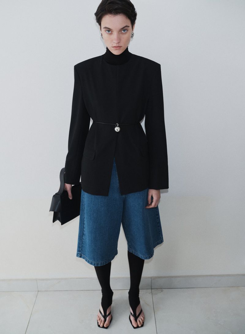 Woman in black jacket, blue denim culottes, black tights, and sandals poses against a white wall, holding a black clutch purse.