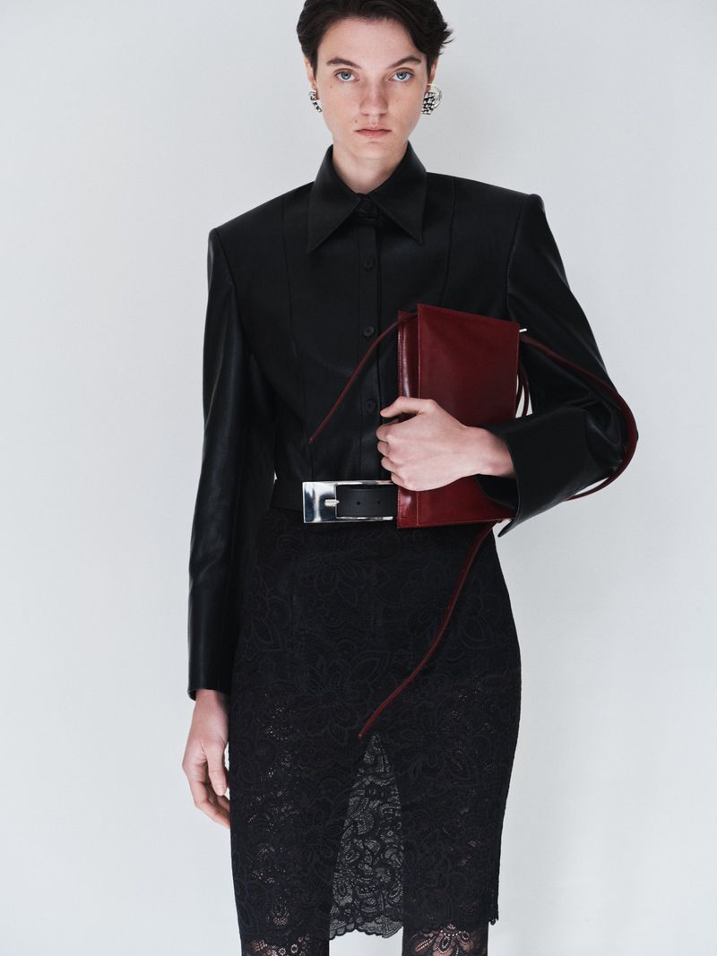 Woman in black lace dress and leather jacket holds a red portfolio. Fashion editorial shot against a white backdrop in studio lighting.