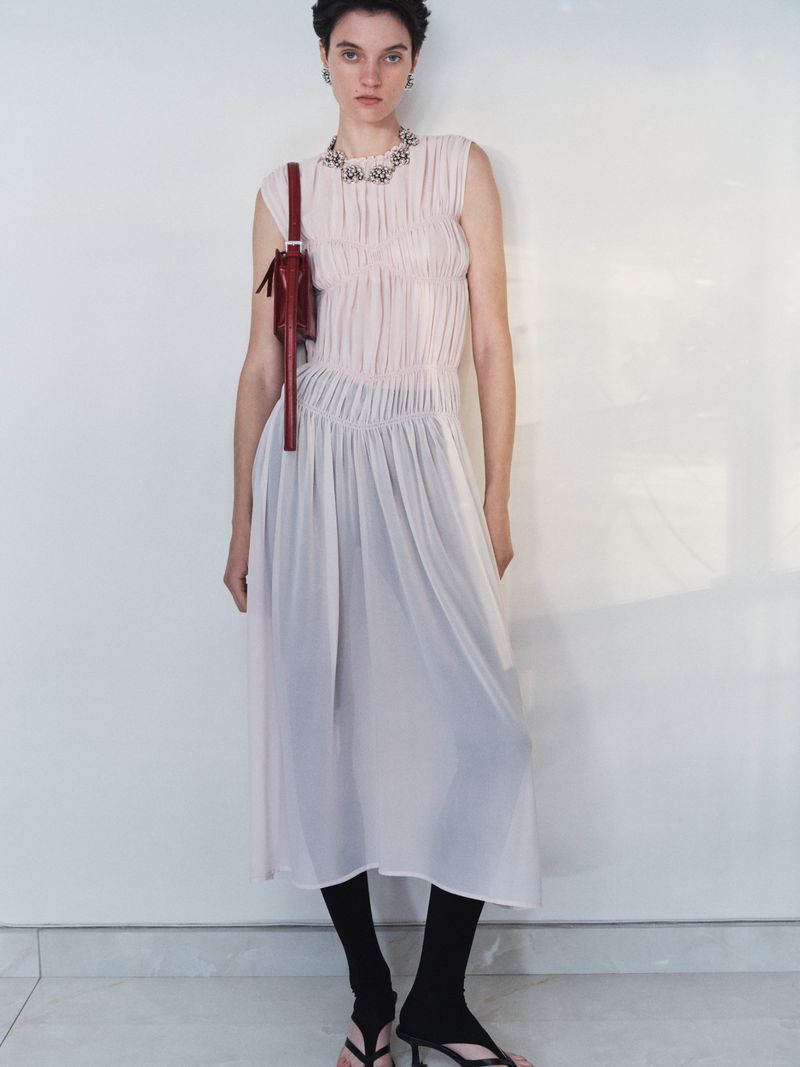Model wearing a pleated pale pink sleeveless dress and carrying a burgundy shoulder bag against a light background