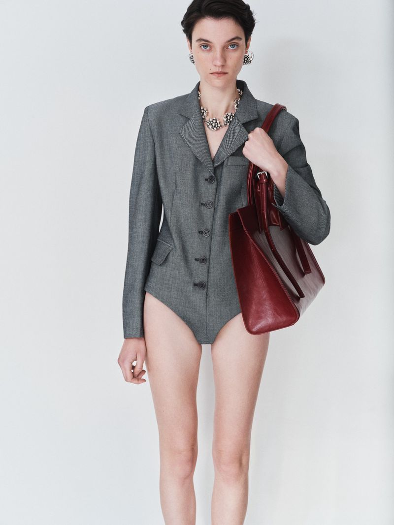 Fashion portrait of a woman with short hair wearing a grey blazer and holding a red tote bag in a white photo studio.