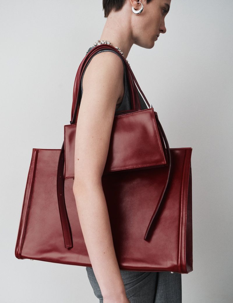 Person carries large burgundy leather tote bag with attached smaller pouch bag in a studio shot against a plain white background.