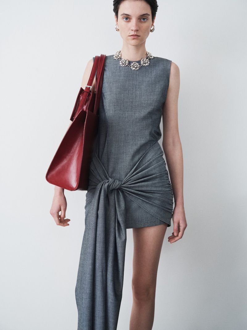Fashion model posing in a gray knotted mini dress with a red tote bag in a photo studio against a neutral backdrop, wearing jewelry.
