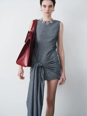 Fashion model posing in a gray knotted mini dress with a red tote bag in a photo studio against a neutral backdrop, wearing jewelry.