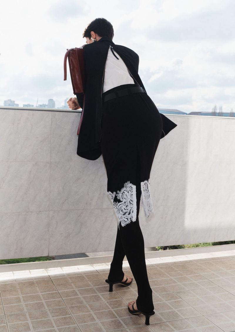 Back view of a woman in black blazer and skirt with lace detail, standing on a terrace overlooking a cityscape on a sunny day.