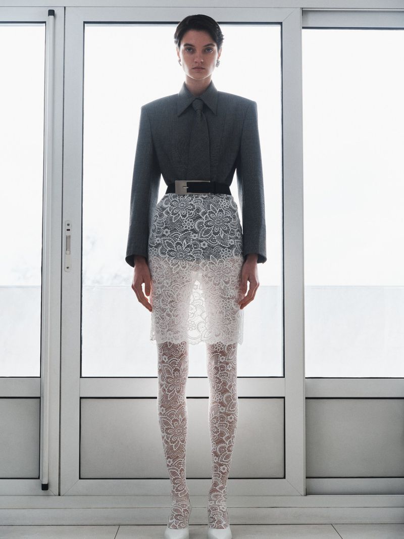A woman in gray blazer, white lace skirt and tights stands in front of bright windows in a fashion forward, full-length shot.