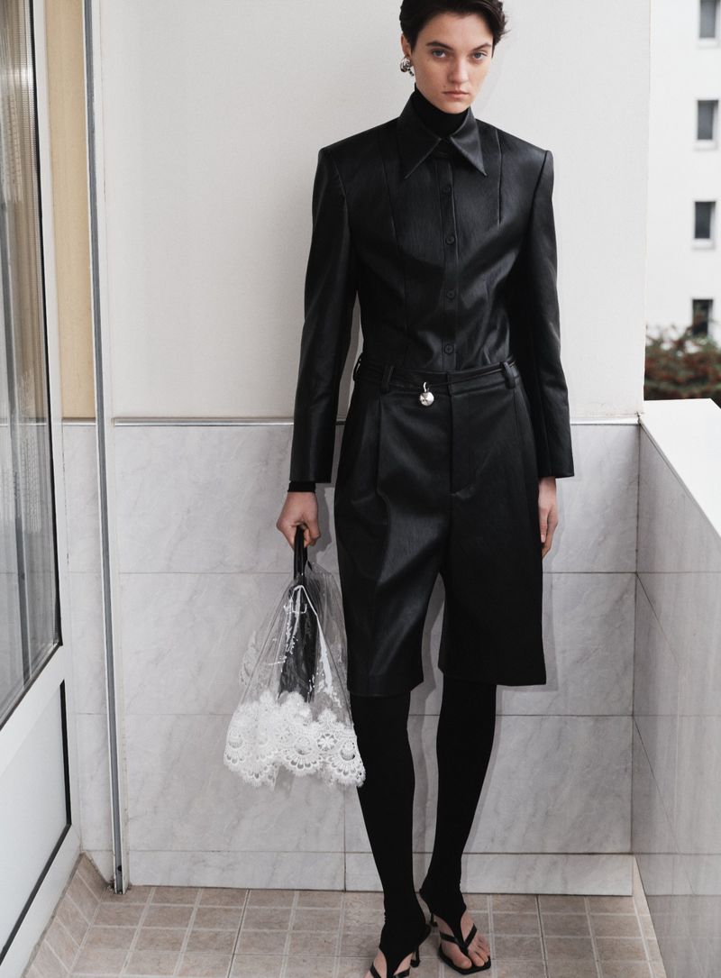 Fashionable woman in a black leather collared jacket, Bermuda shorts, and leggings holds a sheer handbag on a balcony.
