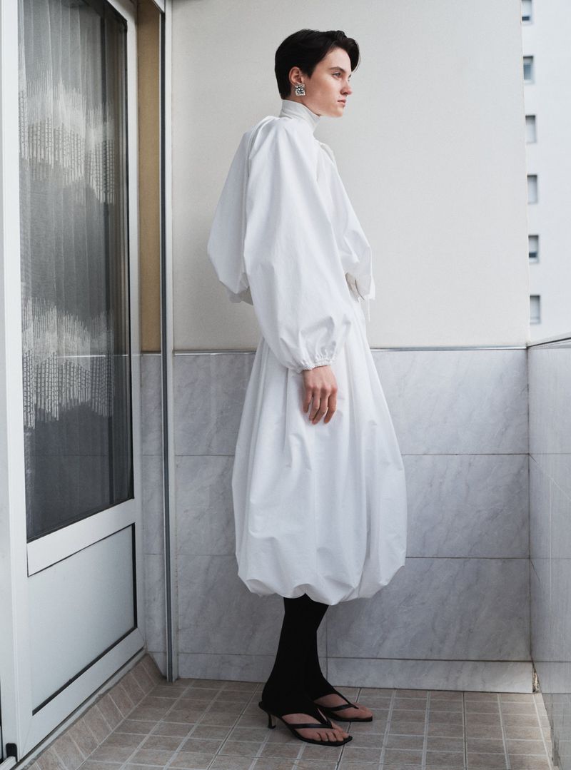 Model standing on a balcony wearing a white voluminous dress and black leggings for Beaufille Pre-Fall 25.