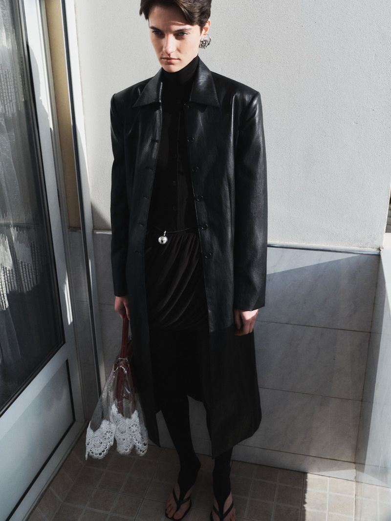 Young woman with short hair wearing a black dress, coat, and flip-flops posing indoors while holding a transparent lace bag.