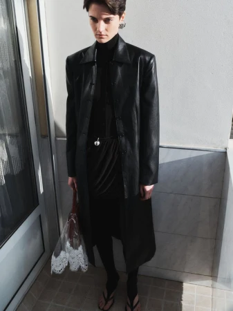 Young woman with short hair wearing a black dress, coat, and flip-flops posing indoors while holding a transparent lace bag.