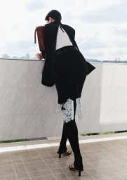 Model in black clothing with white lace detail leaning against a white tiled wall overlooking a city skyline.