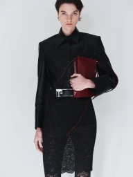 Model in black button-down shirt and lace skirt holding a burgundy handbag against a white studio background.