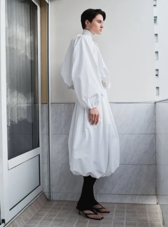 Model standing on a balcony wearing a white voluminous dress and black leggings for Beaufille Pre-Fall 25.