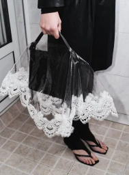 Model wearing black clothing holding a black handbag covered by a clear plastic shield with white lace trim