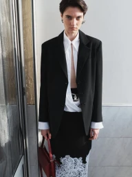 Model in a black blazer and white shirt holding a dark red handbag standing outdoors against a wall