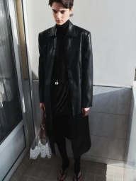 Model wearing a black leather coat and dark turtleneck holding a transparent handbag standing outdoors in bright daylight