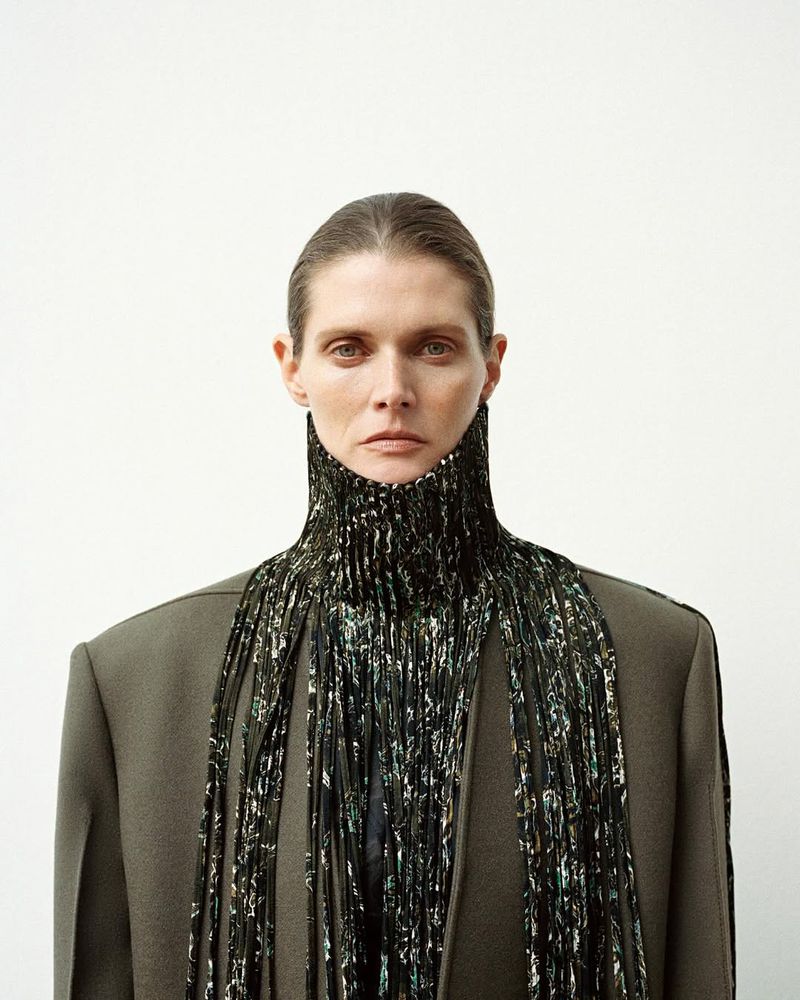 Woman in a grey jacket and a decorative black and silver high collar in a studio setting. Fashion editorial shot.