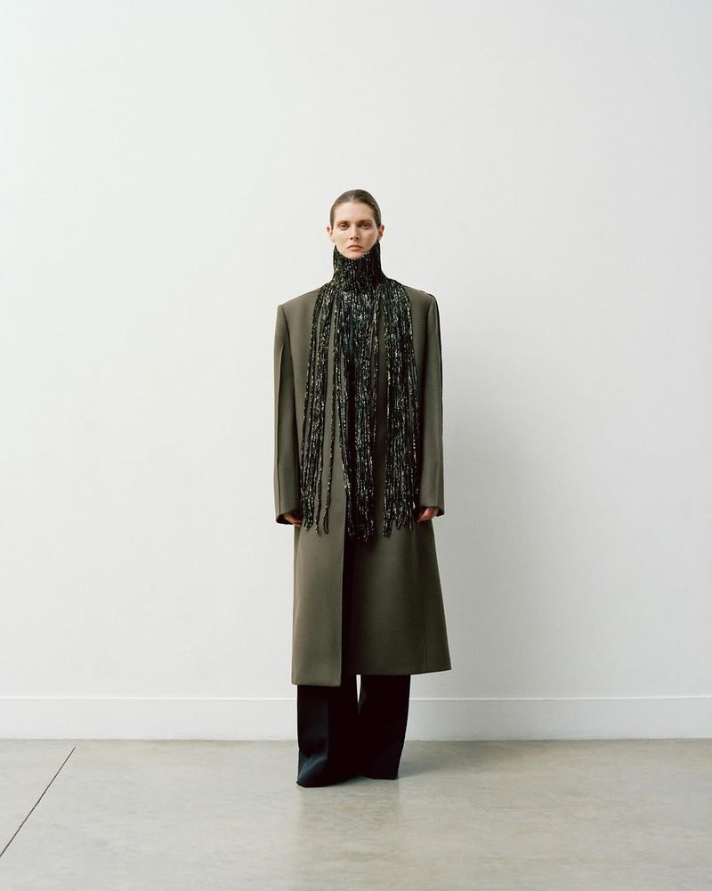Full-length portrait of a fashion model in a long coat with a beaded scarf, standing in a photo studio with a white wall.