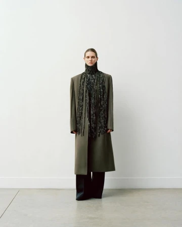 Full-length portrait of a fashion model in a long coat with a beaded scarf, standing in a photo studio with a white wall.