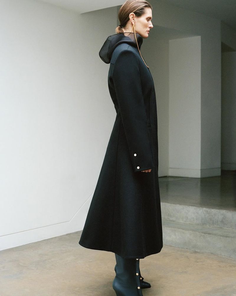 Adult woman in a black hooded coat and heeled boots stands in a minimalist interior setting, showcasing fashion and style.