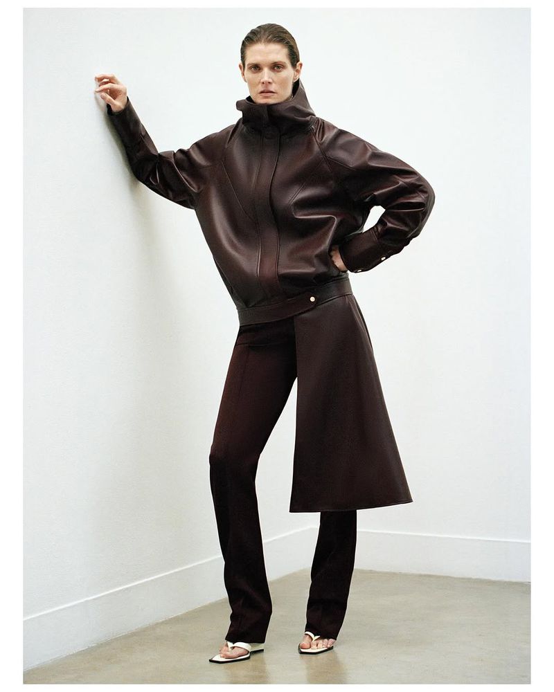 Model Malgosia Bela wearing dark brown leather jacket and matching trousers leaning against a white studio wall