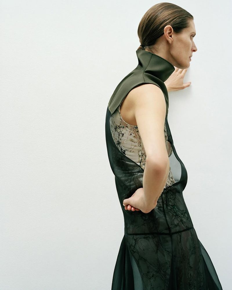 A woman in an olive green collar and semi-sheer black dress poses sideways in front of a plain white wall in a fashion editorial.