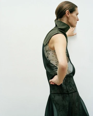 A woman in an olive green collar and semi-sheer black dress poses sideways in front of a plain white wall in a fashion editorial.