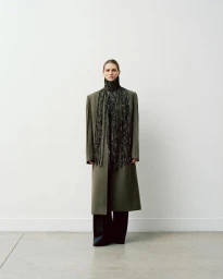 Model wearing a long olive green coat and beaded scarf against a neutral white background