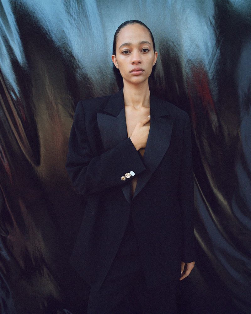 Portrait of a slender Black woman with a neutral expression. She is wearing a black blazer, and there is a silvery, draped background.