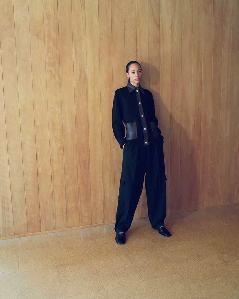 An adult woman poses in a black jacket and pants against a wood wall. She looks directly at the camera in a fashion shot.