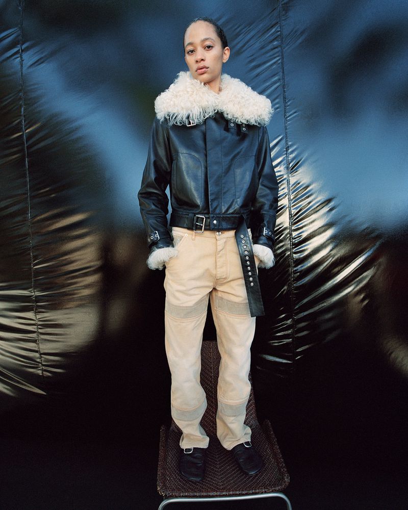 A person poses in a studio wearing a black leather bomber jacket with cream-colored pants and black boots against a dark background.