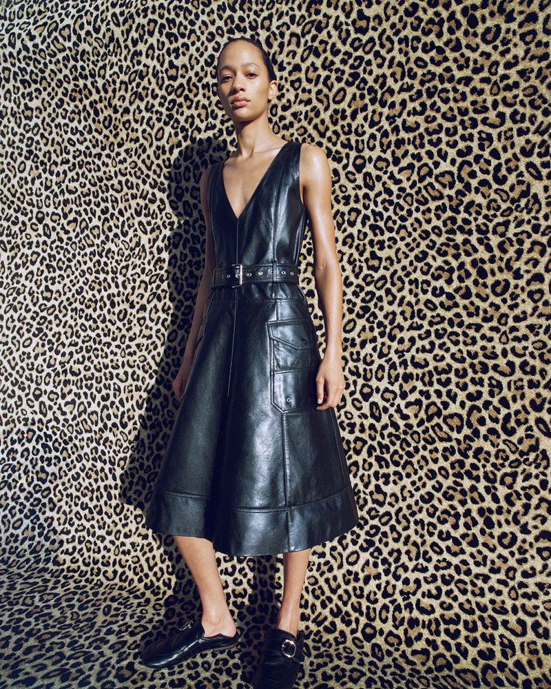 A woman poses in a sleeveless, knee-length, black leather dress with a belt and flat shoes, standing in front of a leopard-print backdrop.