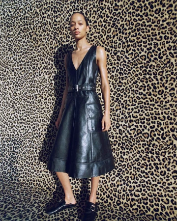 A woman poses in a sleeveless, knee-length, black leather dress with a belt and flat shoes, standing in front of a leopard-print backdrop.