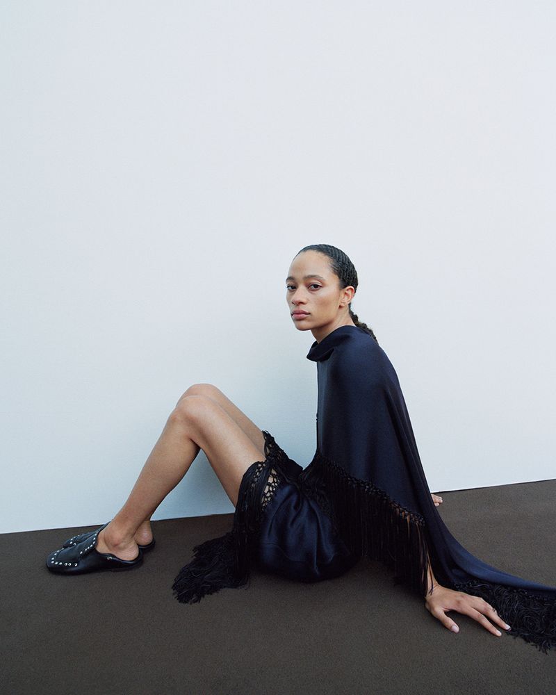 A woman sits in a dark blue dress with fringe, facing the camera in a studio with a light background, wearing black studded flats.