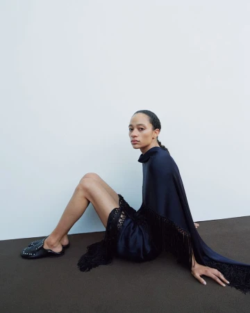 A woman sits in a dark blue dress with fringe, facing the camera in a studio with a light background, wearing black studded flats.