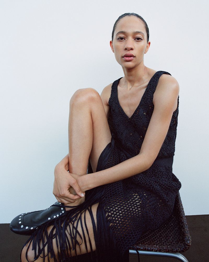 Model Selena Forrest wearing a black fringed knit dress and studded black shoes against a plain white background