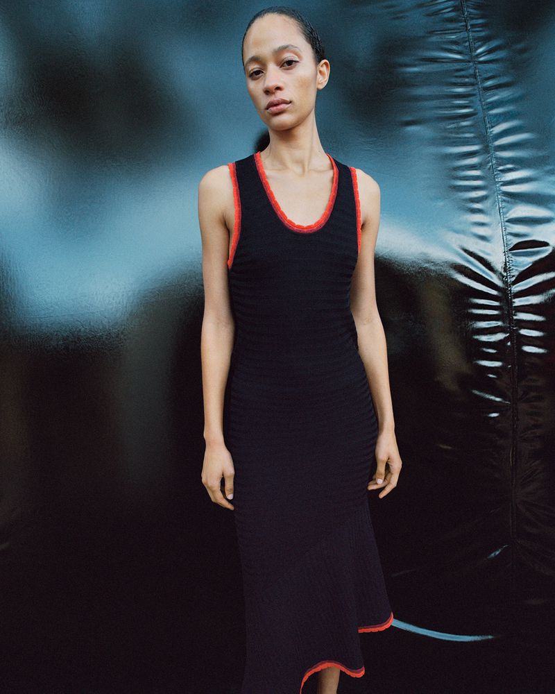 Elegant woman in a black dress with orange trim posing in front of a dark, textured backdrop in a fashion photography setting.